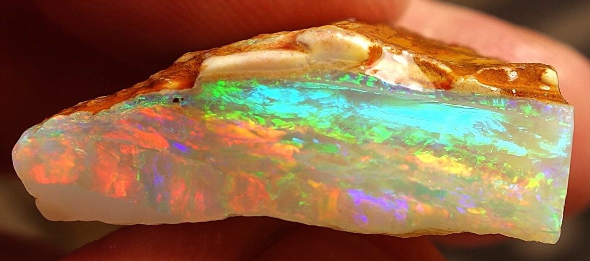 A rough opal specimen from Coober Pedy, South Australia, held between two fingers and showing bands of green, blue, red, and violet play-of-color against its pale matrix