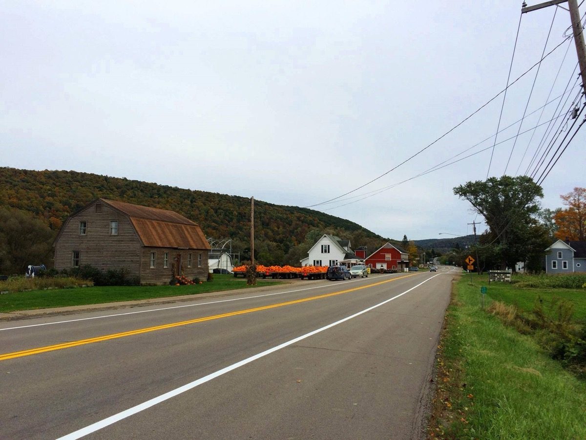 A rural road in Great Valley, New York in October, lined with a weathered barn, pumpkins stacked for sale, and hillsides turning autumn color